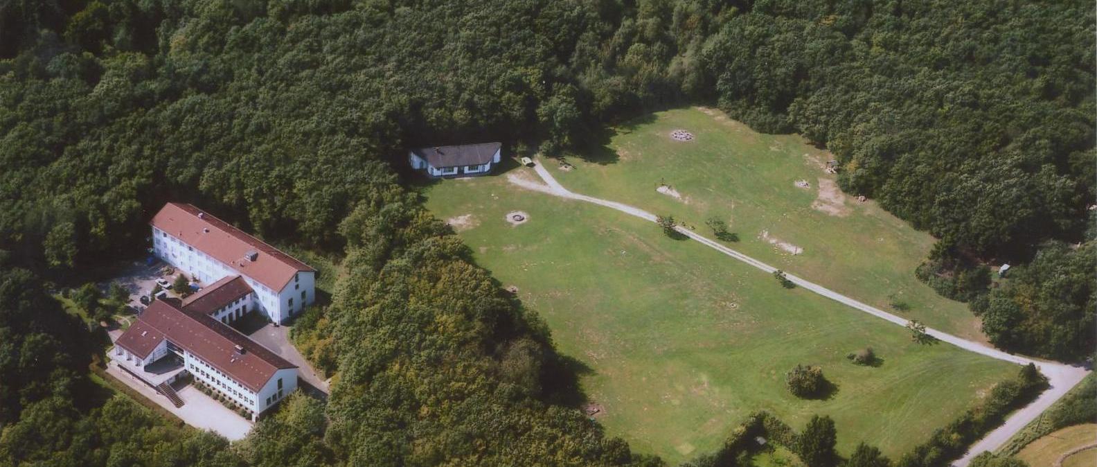 Ansicht des Geländes der Bildungsstätte Steinbachtalsperre. Das Hauptgeäude befindet sich mittig im linken Drittel der Aufnahme, umgeben von Wald, auf der rechten Seite befindet sich eine große Lichtung mit einem Nebengebäude am oberen Rand der Lichtung.