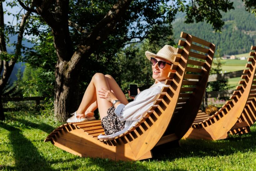 Eine Frau sitzt auf einer Outdoor Sitzgelegenheit aus Holz auf einer Wiese vor einem Baum und einem Busch